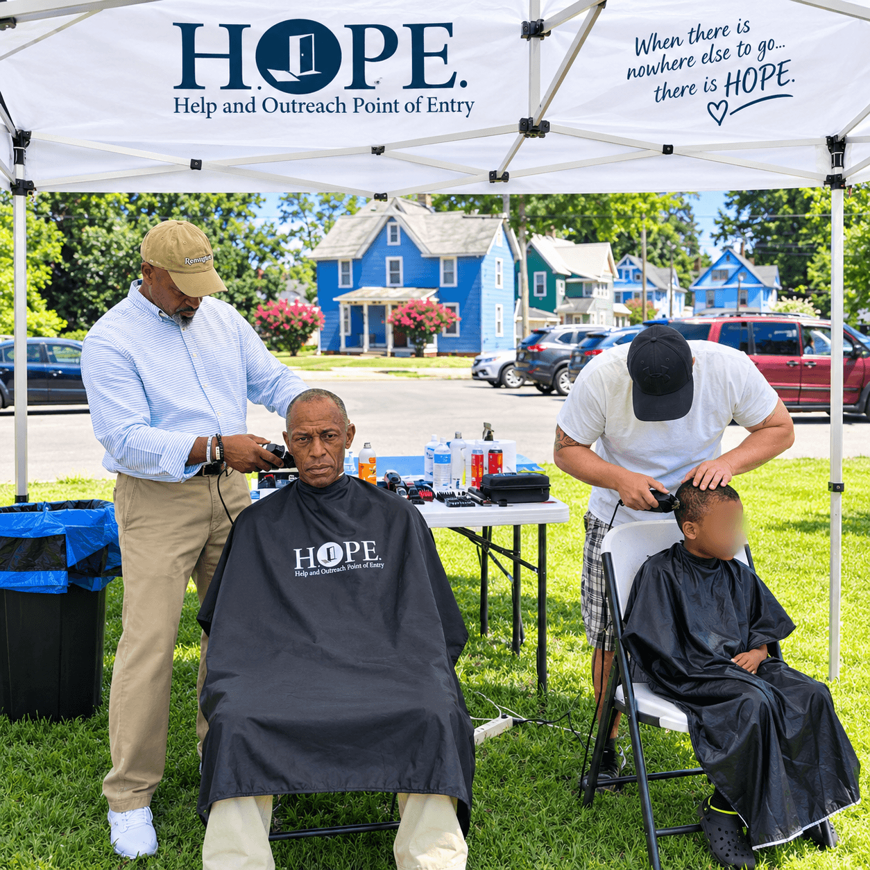 Two men give free haircuts to children under a HOPE tent outdoors, with colorful houses in the background.