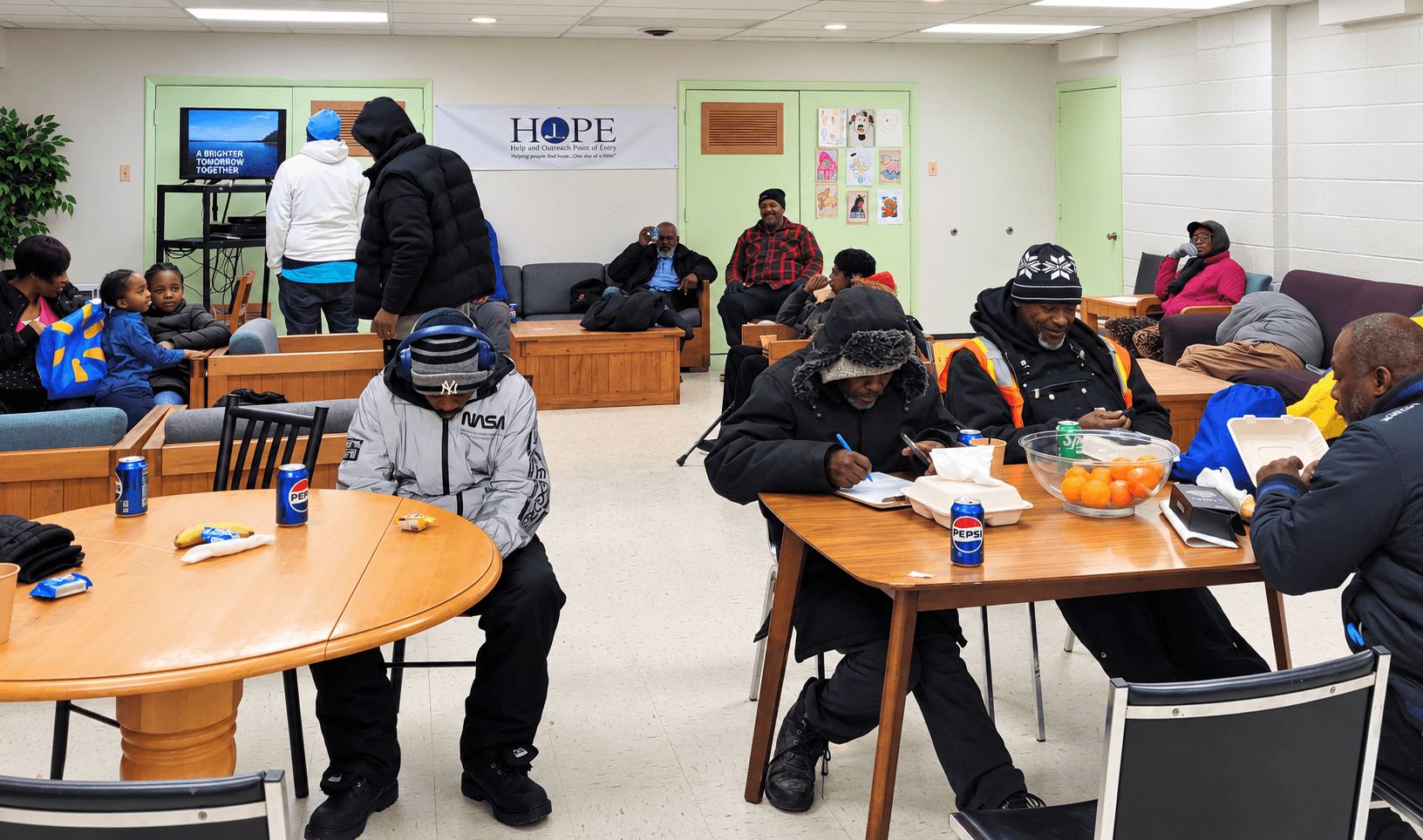 People sitting and standing in a community room with tables, chairs, and a TV, engaging in various activities.
