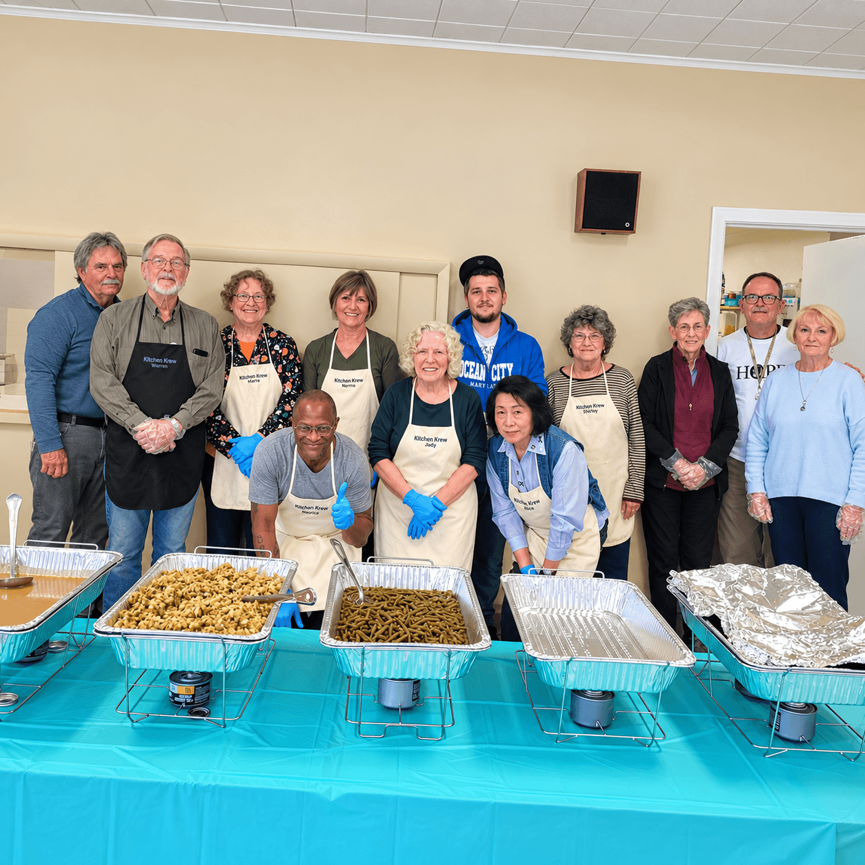 Group of people standing behind a table with food trays, some wearing aprons, in a room with beige walls.