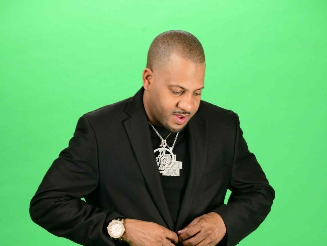 Man with short hair wearing a black suit and jewelry, looking down, against a green background.