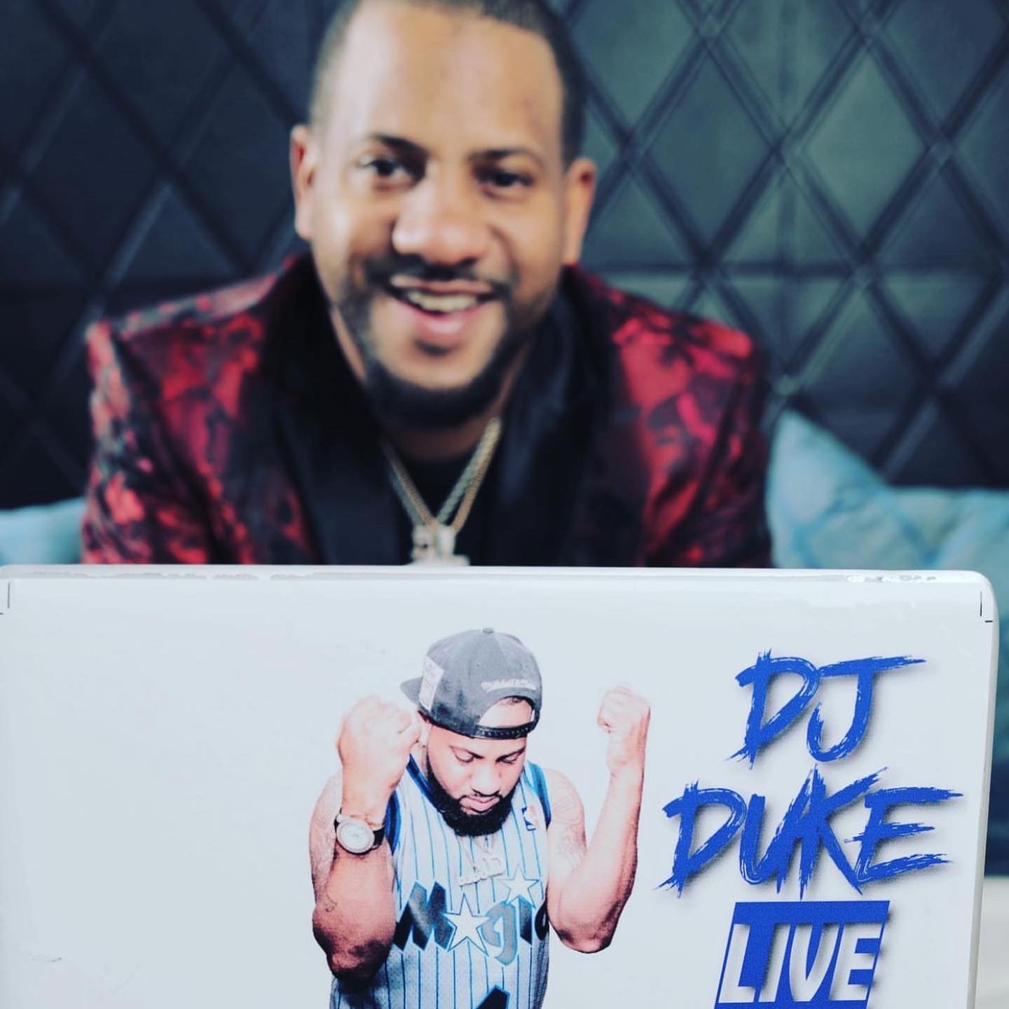 Person smiling behind a sign with a muscular figure, DJ DUKE LIVE text, dark background.