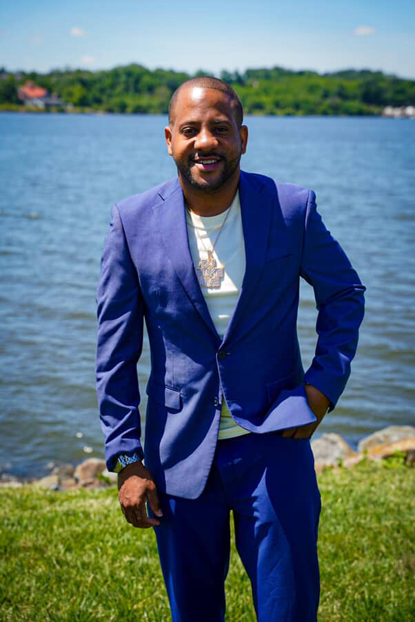 Man in blue suit standing outdoors near water with trees in background, smiling, holding papers in his left hand.