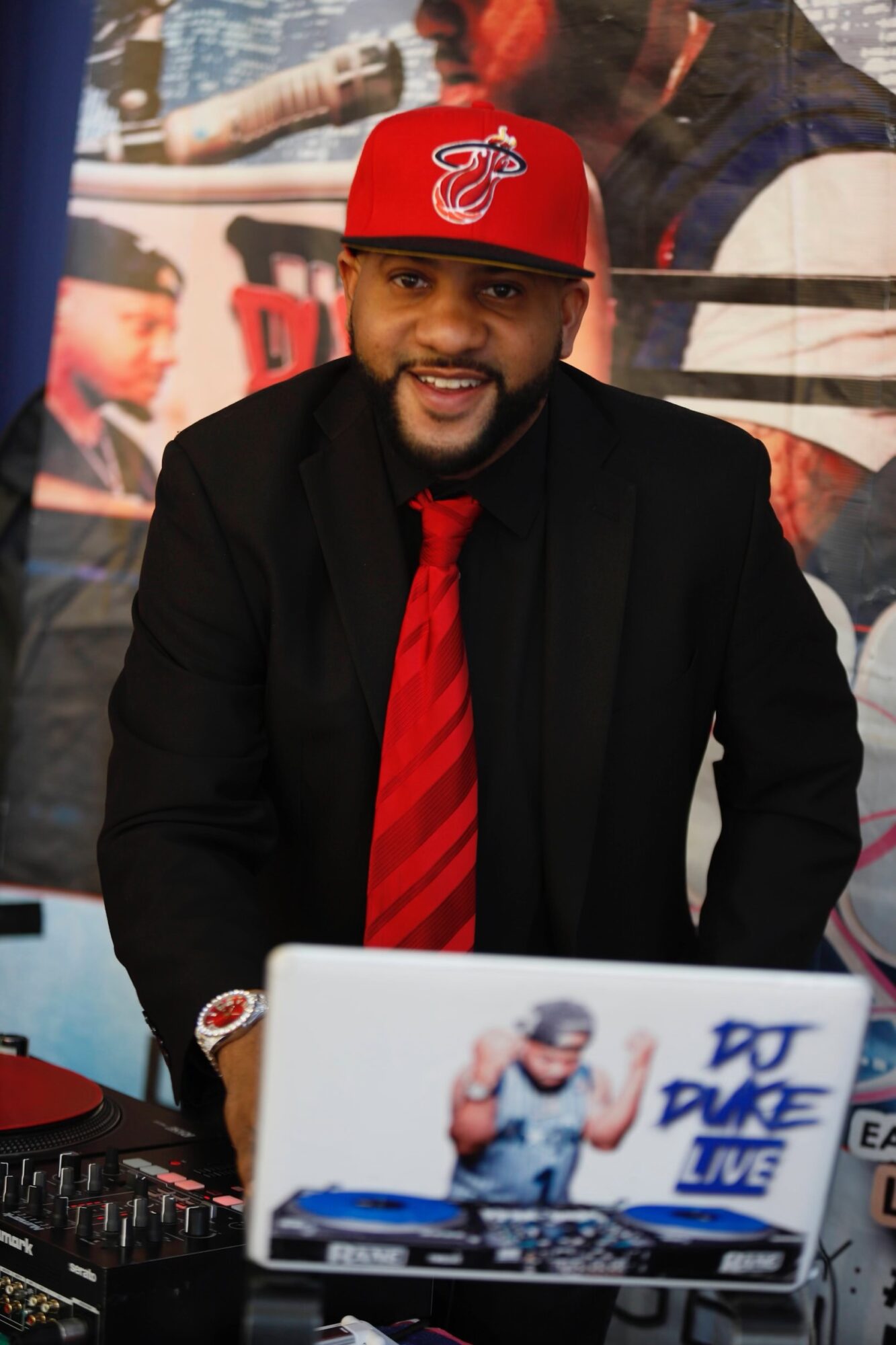 Man wearing a red cap, black suit, and red tie, smiling at a DJ setup with a laptop and equipment.