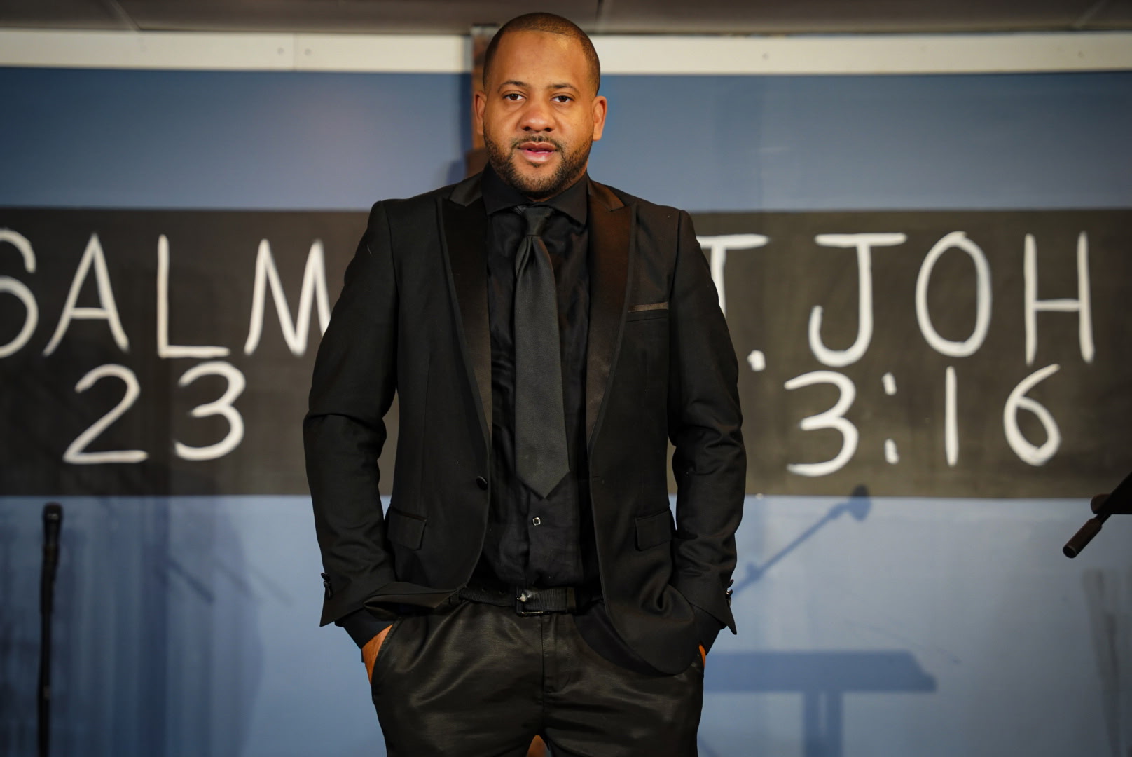 Man in black suit standing in front of a blackboard with white chalk writing, hands in pockets, smiling.