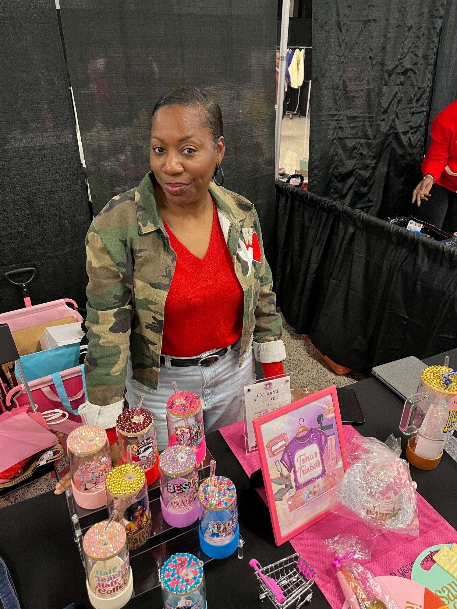Woman standing behind a table with colorful bottles and gift bags at a market or event.