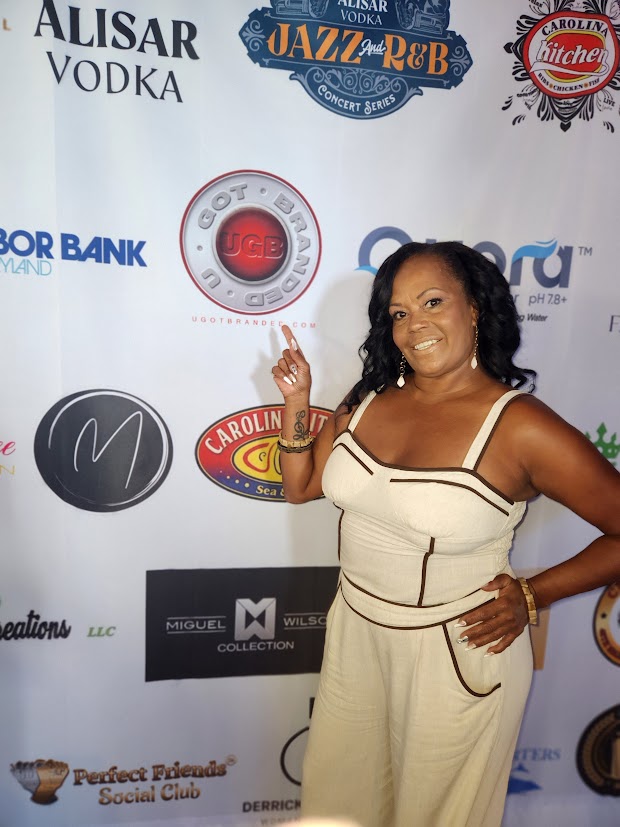 Woman in a cream dress smiling and pointing upward, standing in front of a promotional backdrop with various logos.