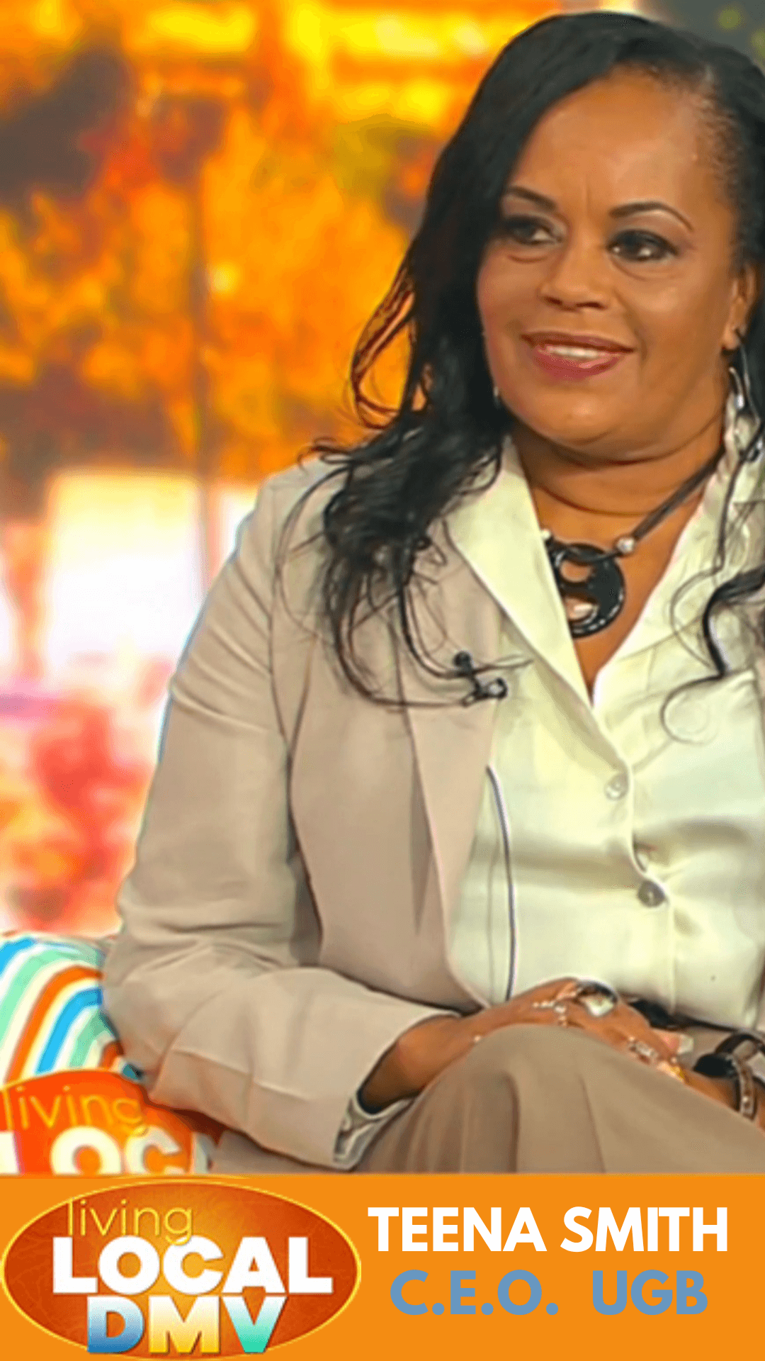 Woman with black hair wearing a beige blazer and cream blouse, sitting outdoors with autumn trees in background.