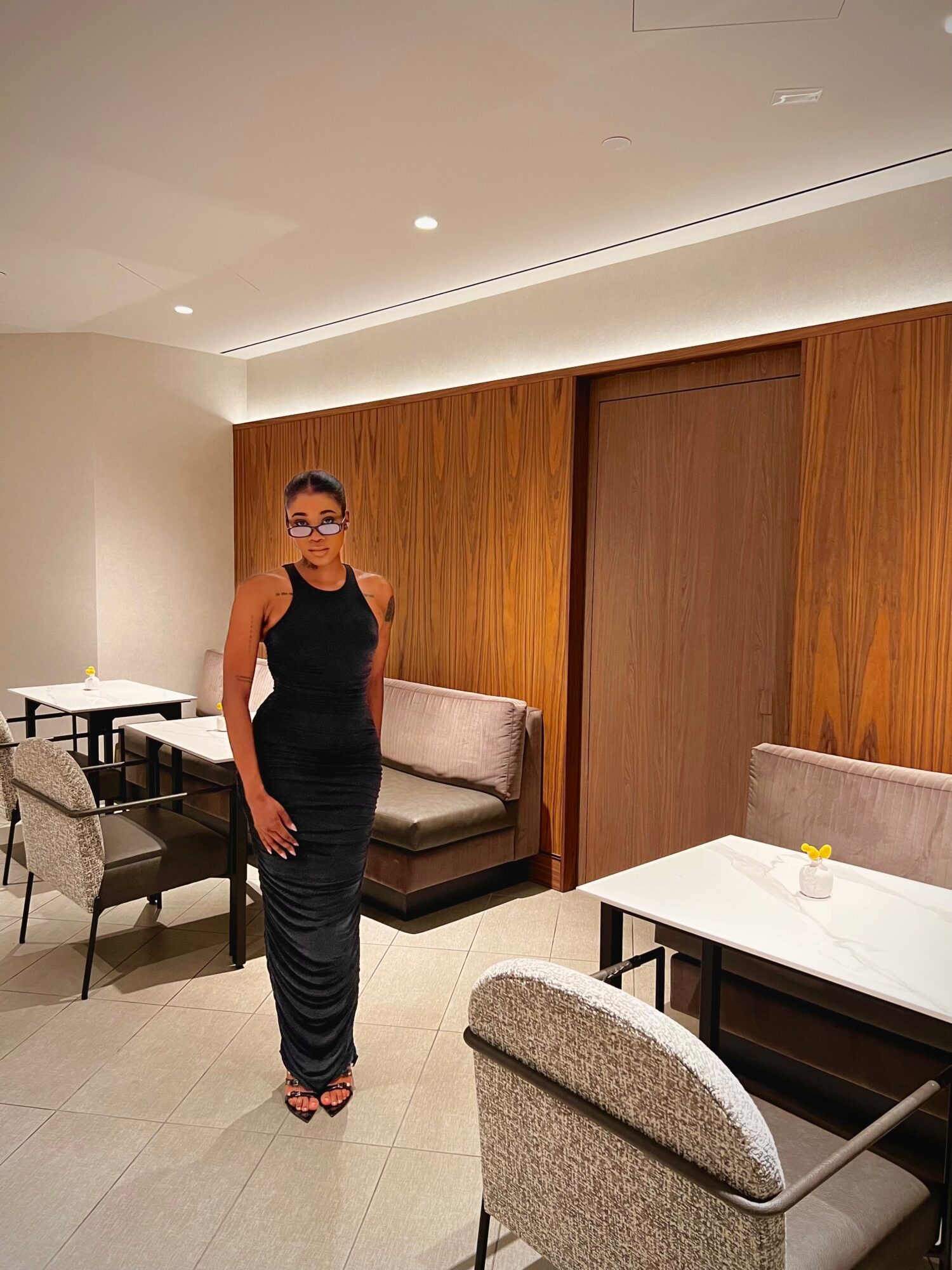 Woman in black dress and glasses standing in a modern room with wooden wall panels and white tables.