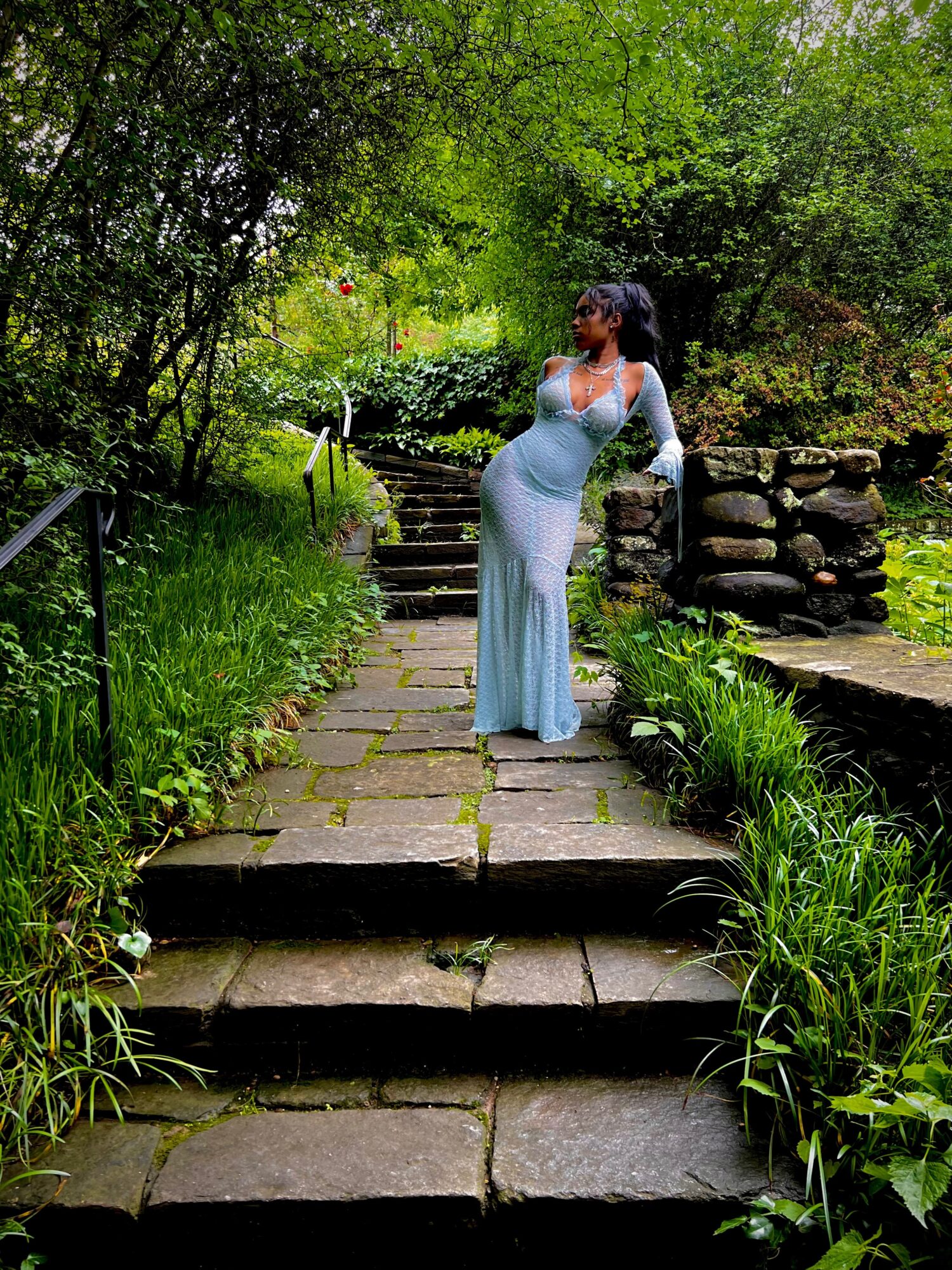 Woman in a long, light blue dress standing on a stone pathway in a lush green garden, surrounded by trees and plants.