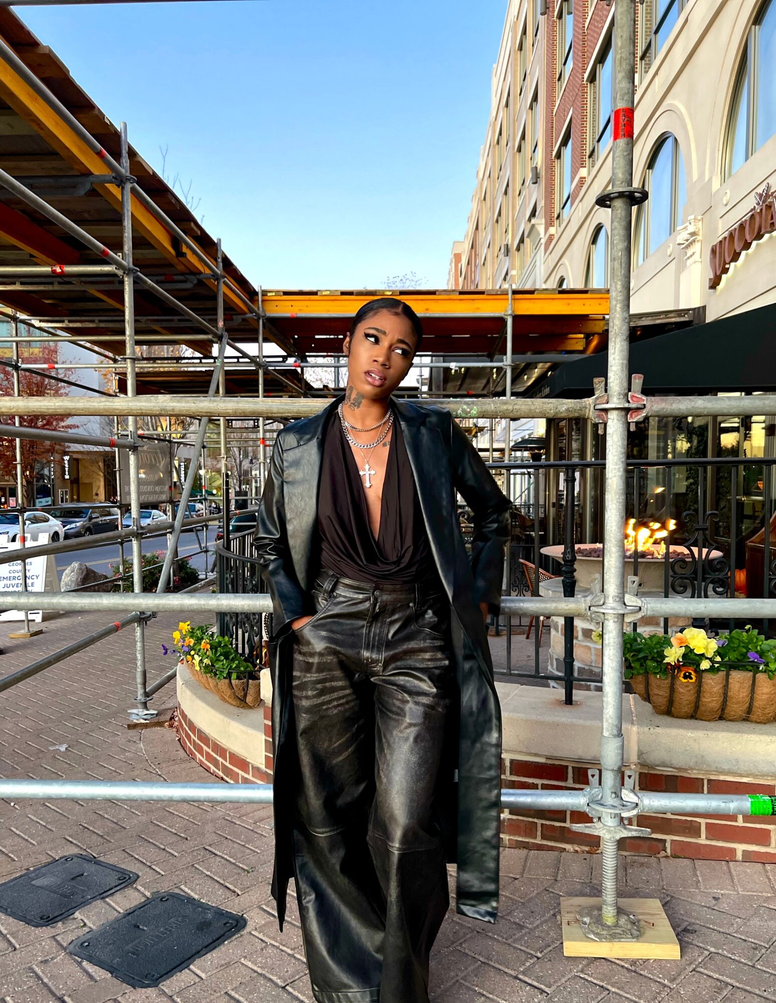 Woman in black leather outfit standing outdoors with scaffolding and buildings in background.