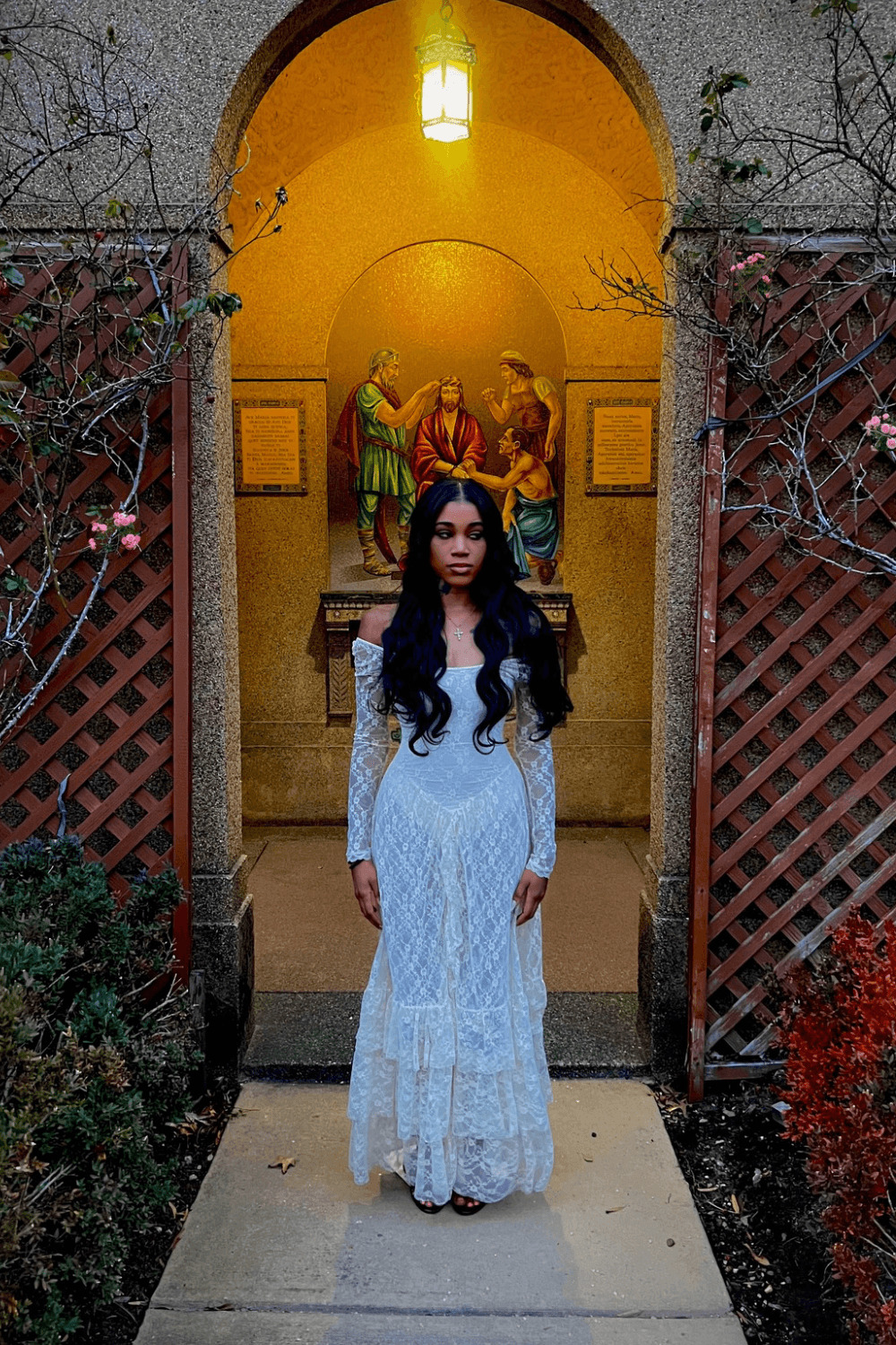 Young woman with long dark hair in a white dress standing in front of a religious statue inside an arched doorway.