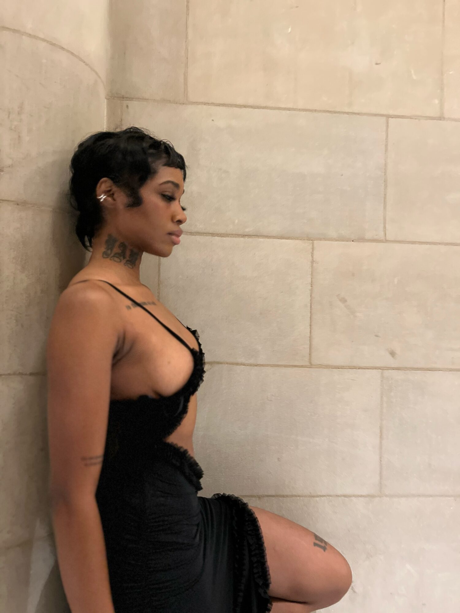 Young woman with short dark hair and tattoos, wearing a black dress, leaning against a beige tiled wall.