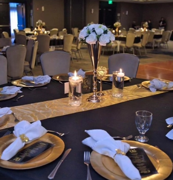 Elegant banquet table with floral centerpiece, candles, gold and black tableware, and neatly folded napkins in a dimly lit event hall.
