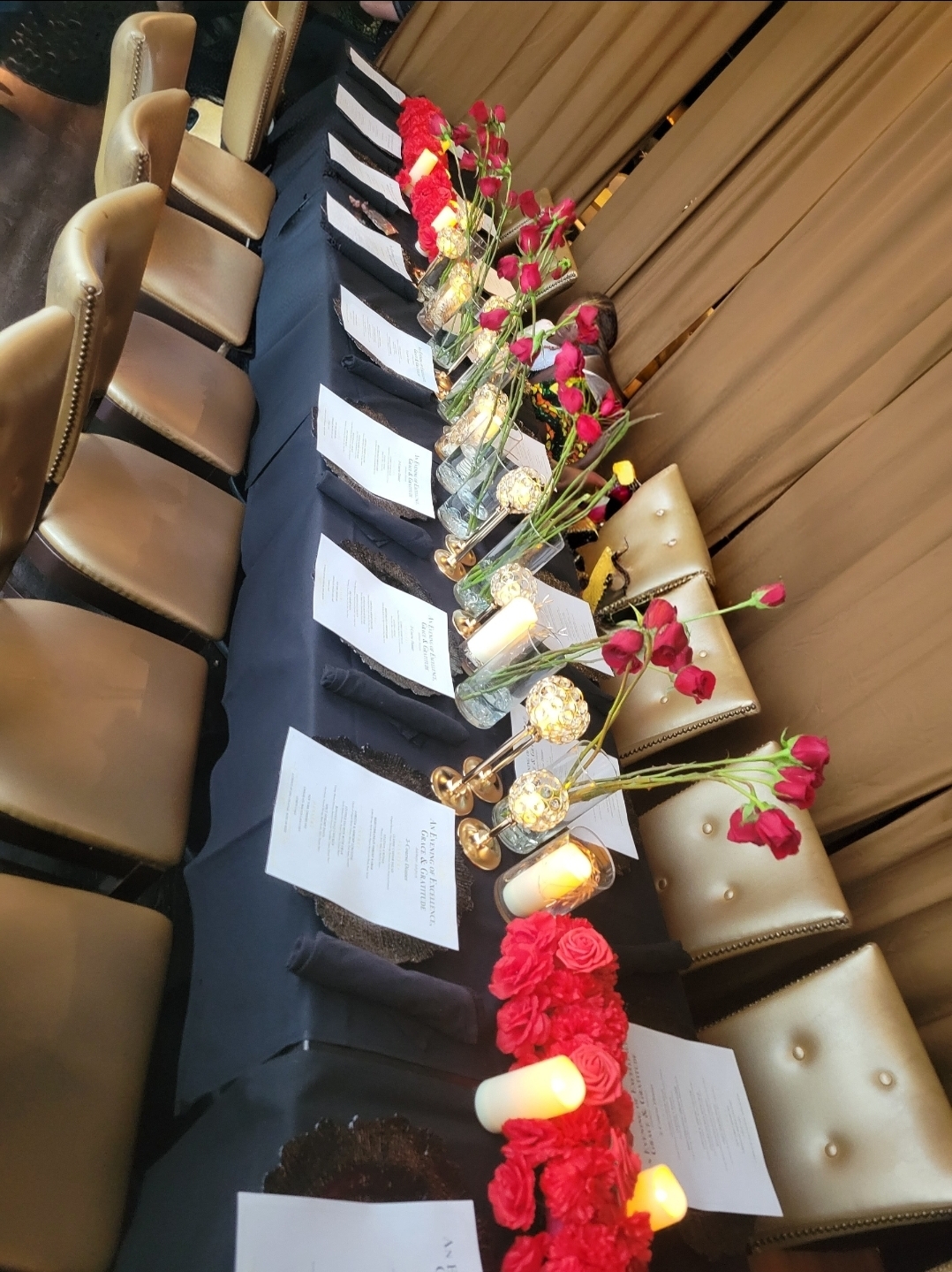 Long table with floral arrangements, candles, and papers, surrounded by chairs, set for an event, with a wooden wall background.