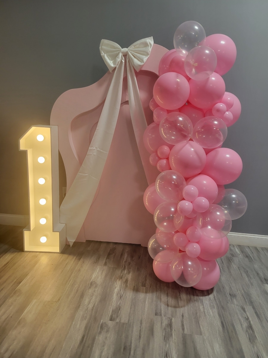 Decorative setup with a large pink gift box, a cream bow, and a cluster of pink and clear balloons, with a lit number one sign.