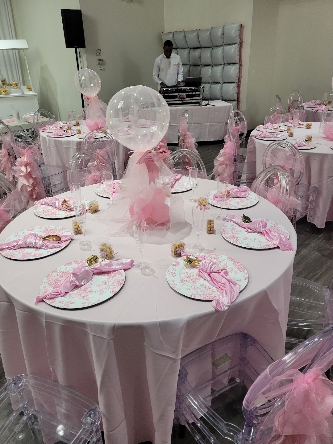Round table decorated with pink and white themed party setup, transparent chairs, balloons, and tableware, in a decorated event space.