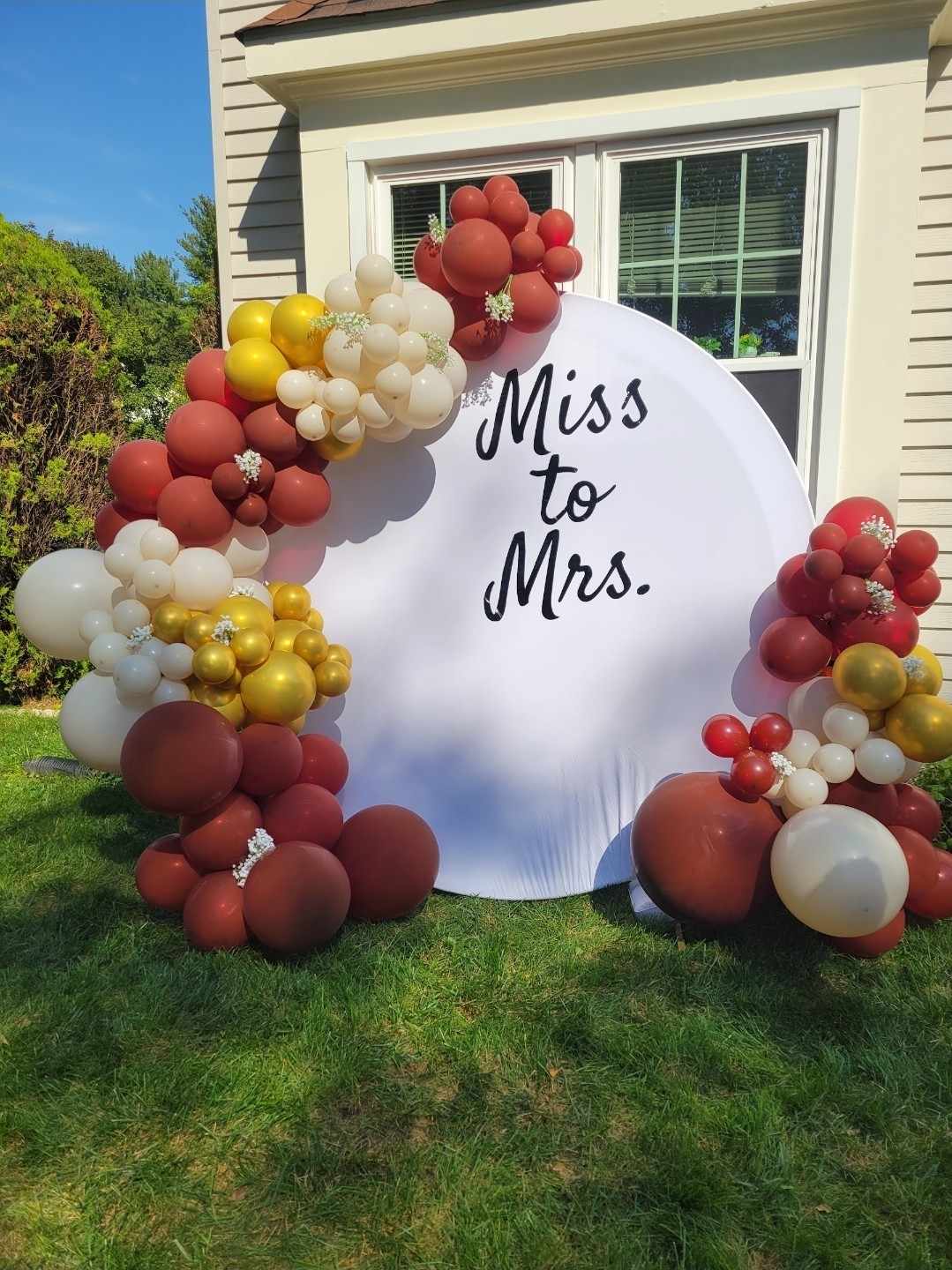 Decorative balloon arch with red, white, yellow, and gold balloons surrounds a white circular sign reading 'Miss to Mrs.' outside a house.