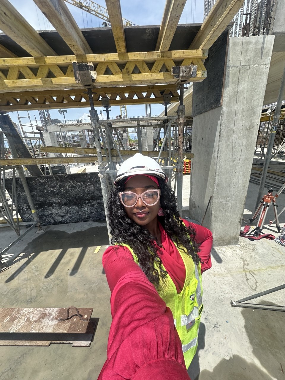 Woman in safety gear taking selfie at construction site with scaffolding and concrete structure behind her.