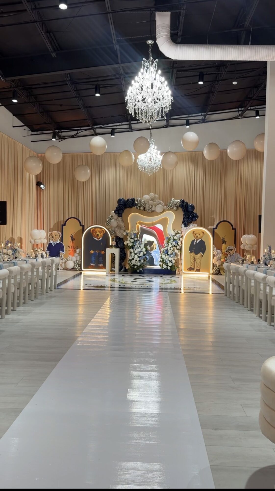 Elegant event hall with a chandelier, decorated stage, and rows of tables and chairs, ready for a celebration.