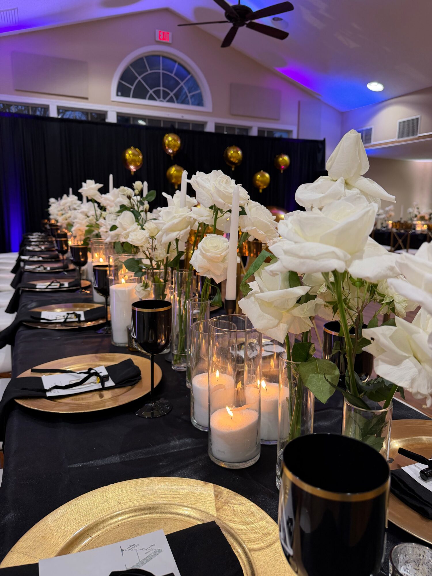 Table with white floral centerpiece, black and gold accents, and candles, set for a formal event.