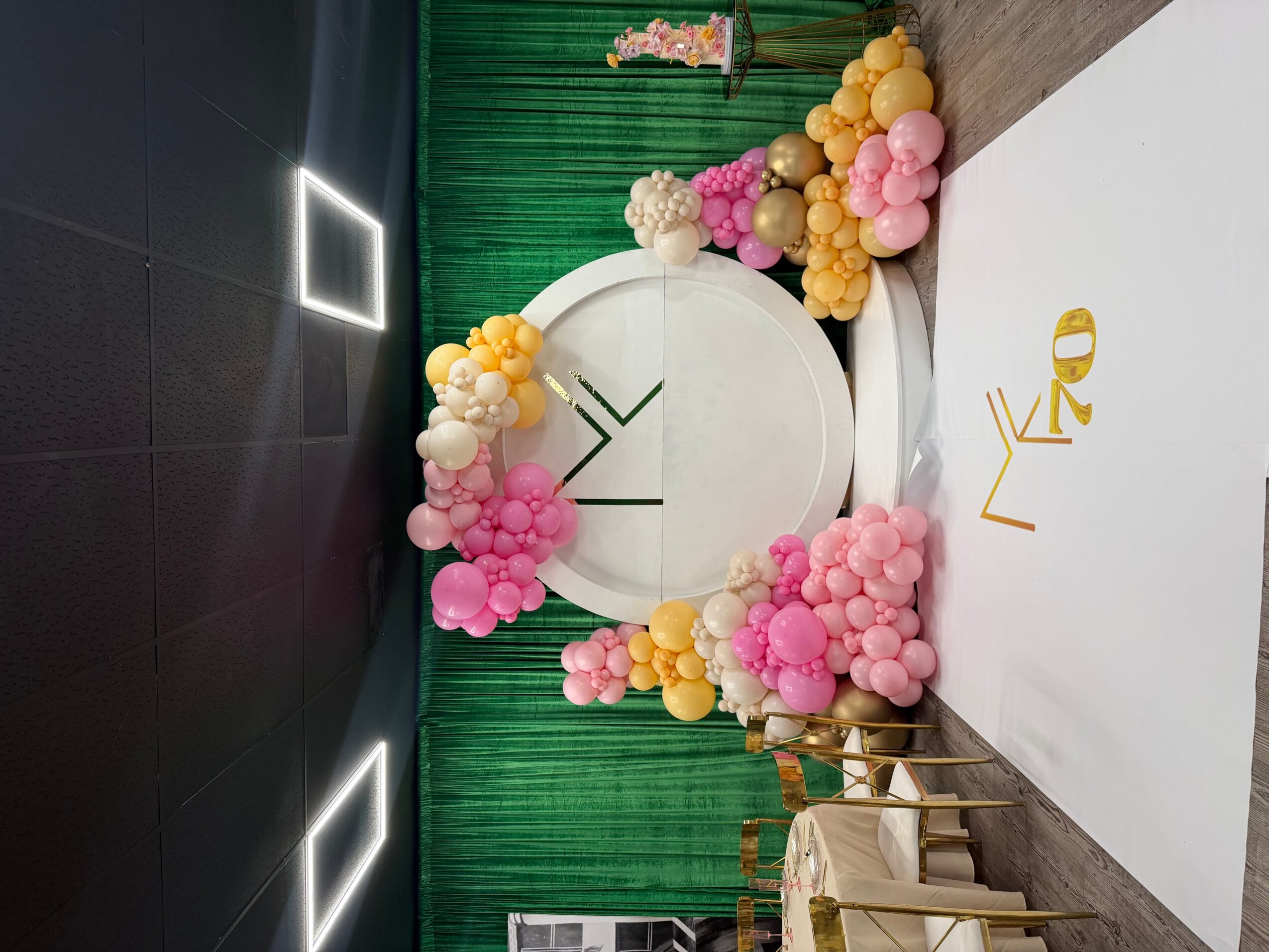 Decorative backdrop with pink, yellow, and white balloons, green curtain, and a white circular sign with black arrow, for celebration.