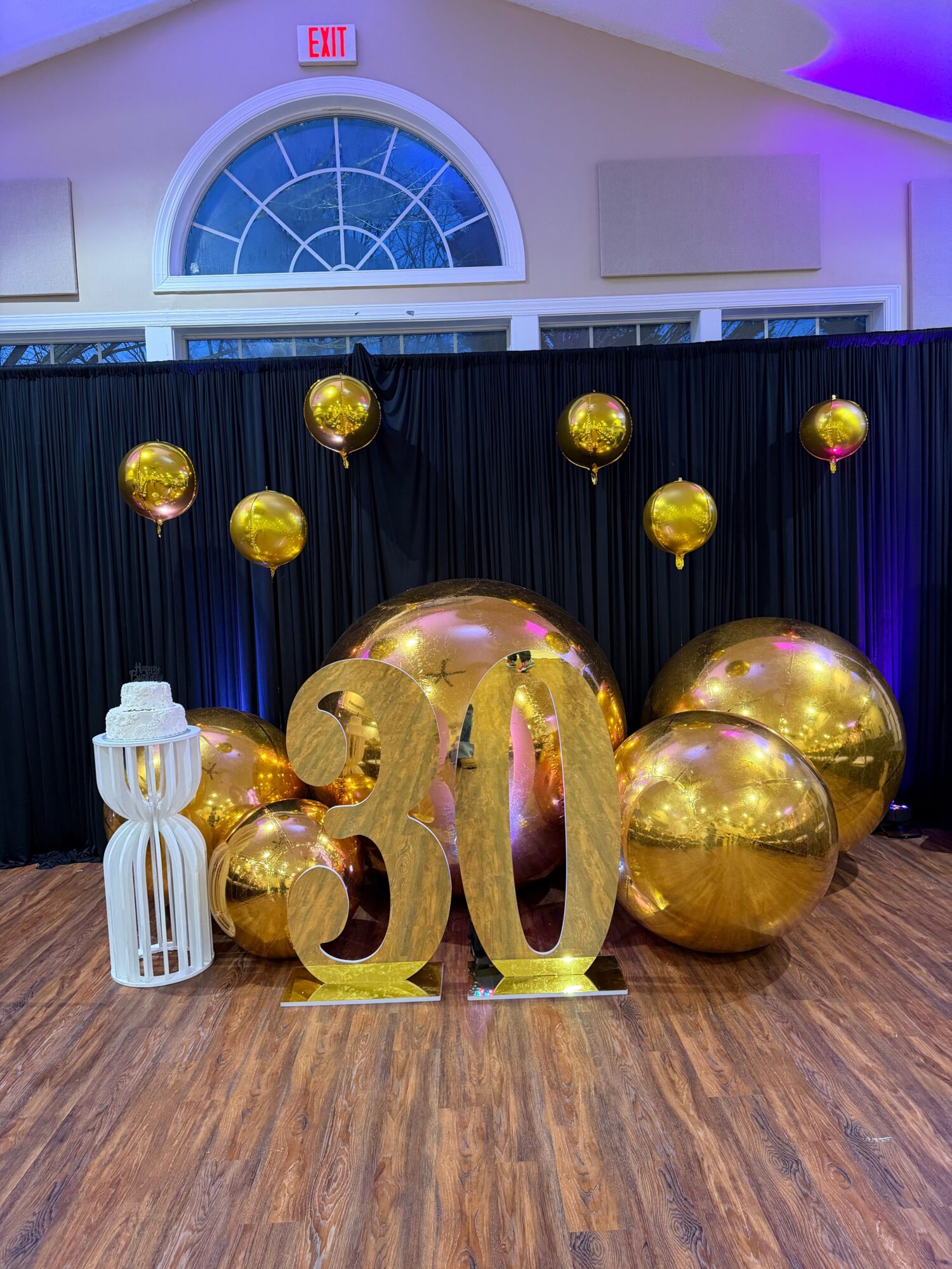 Gold balloons of various sizes arranged on a wooden floor with a black curtain background and a white decorative object.