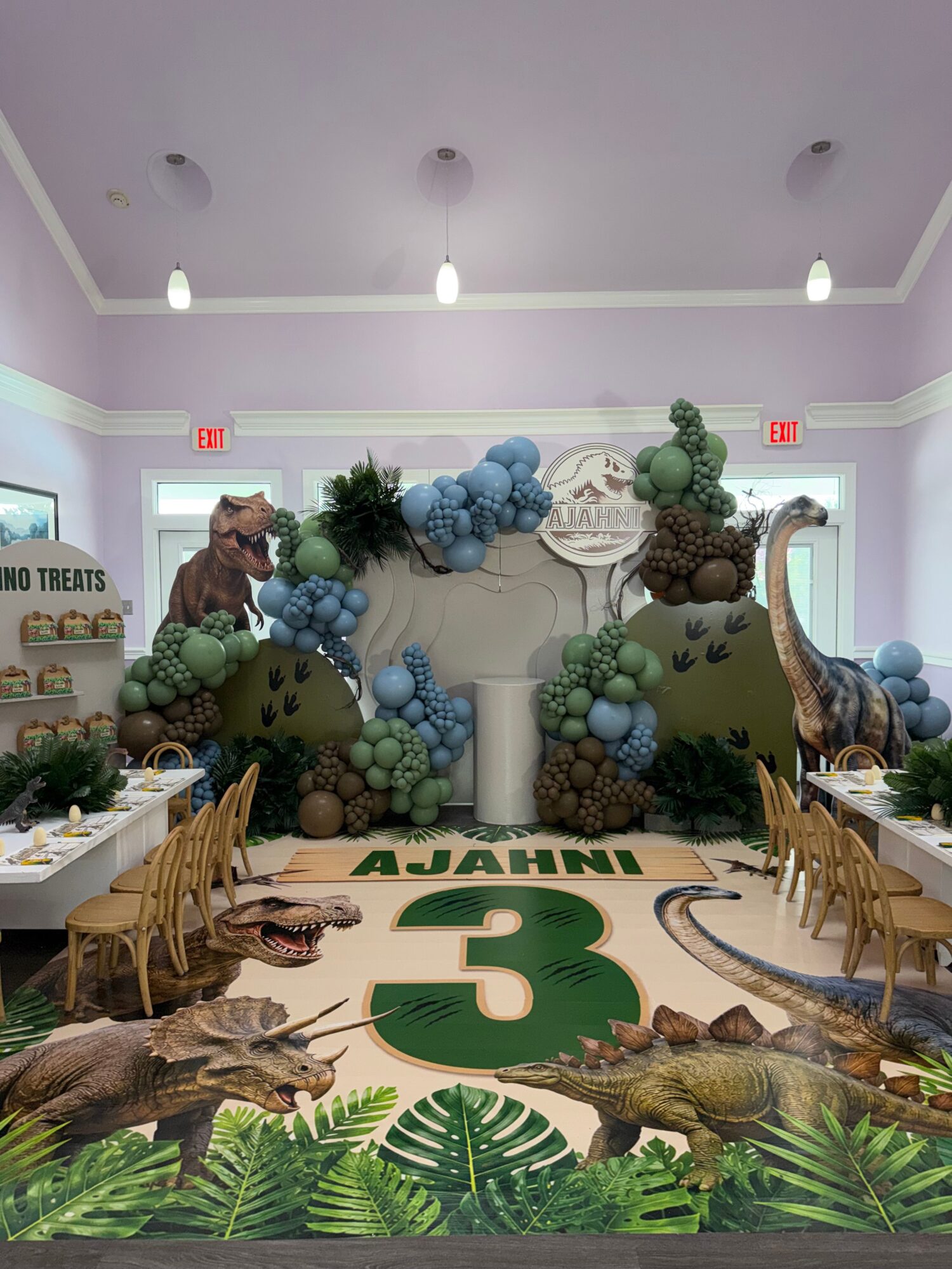 Party setup with dinosaur decorations, balloons, and a jungle-themed backdrop, featuring a large number three and green foliage.