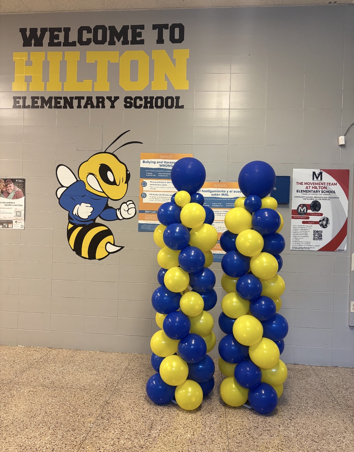 Balloon columns with blue and yellow balloons in front of a wall with a bee mascot and welcome sign at Hilton Elementary School.