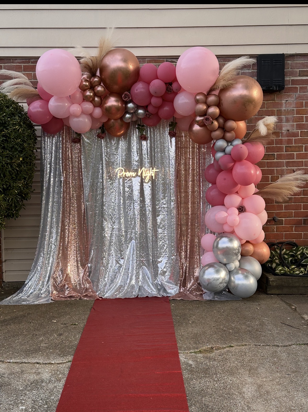 Balloon arch with pink, bronze, and silver balloons, backdrop with silver curtains, red carpet, and brick wall background.