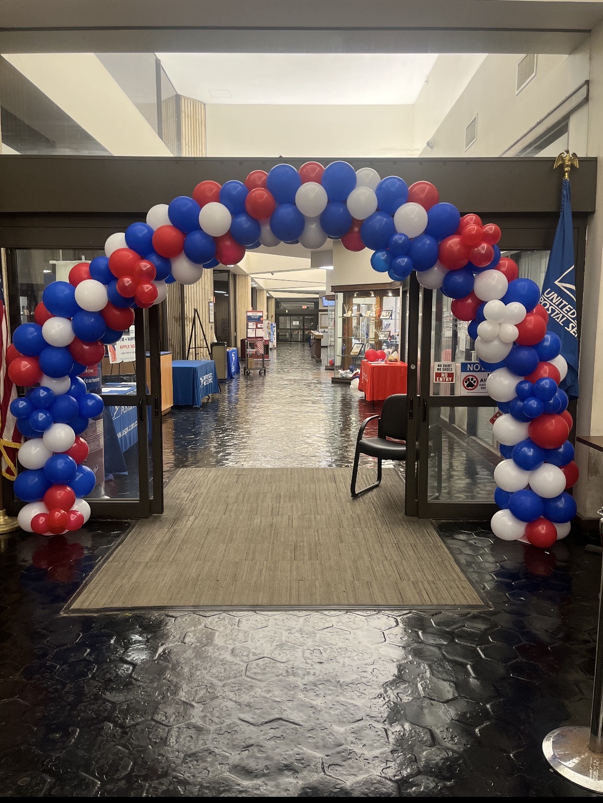 Balloon arch with red, white, and blue balloons at entrance of indoor space, leading to a hallway with chairs and tables.
