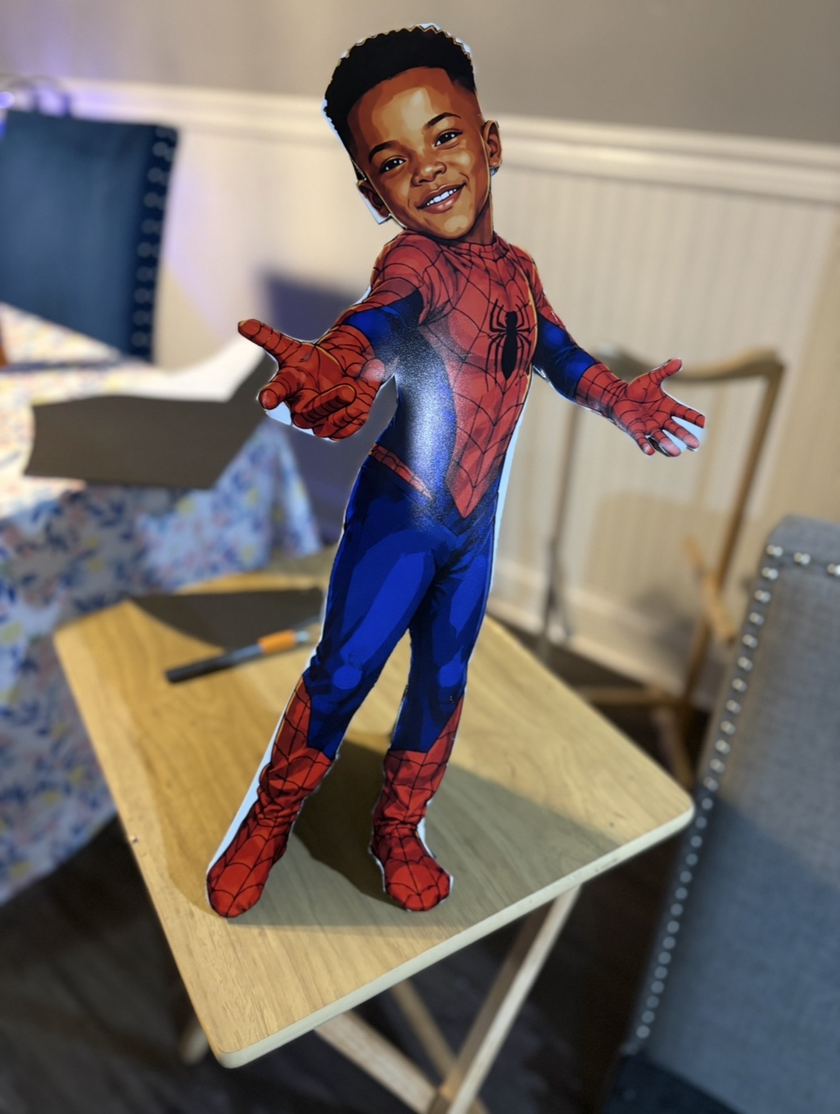 Child dressed as Spider-Man standing on a table, smiling and reaching forward with one hand. Indoor setting with chairs and table.