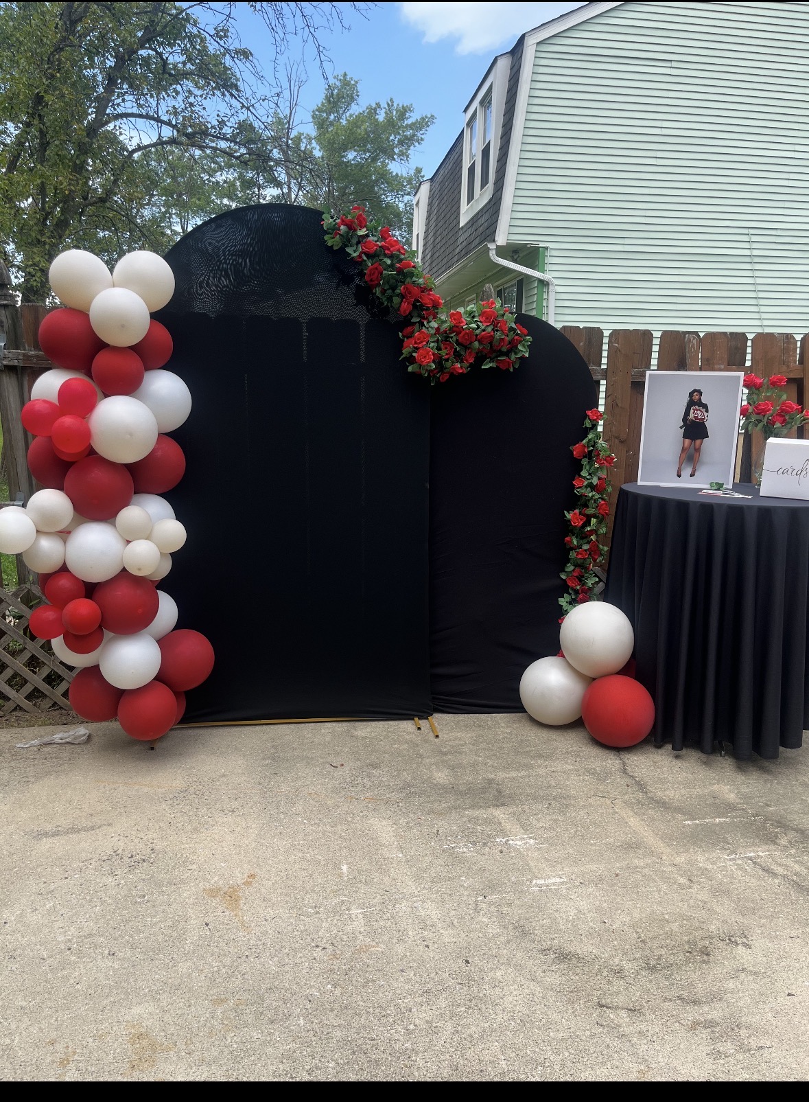 Black backdrop decorated with red and white balloons and red flowers, with a table and framed photo nearby.