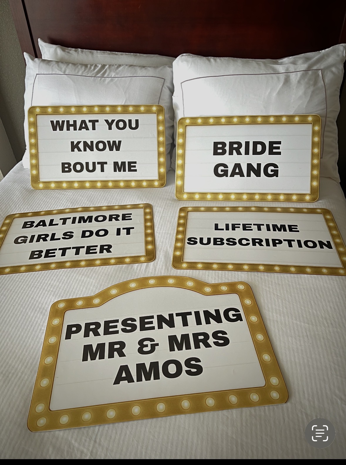 Six decorative signs with bold text on a bed, including messages about a bride gang, Baltimore girls, lifetime subscription, and presenting Mr. and Mrs. Amos.