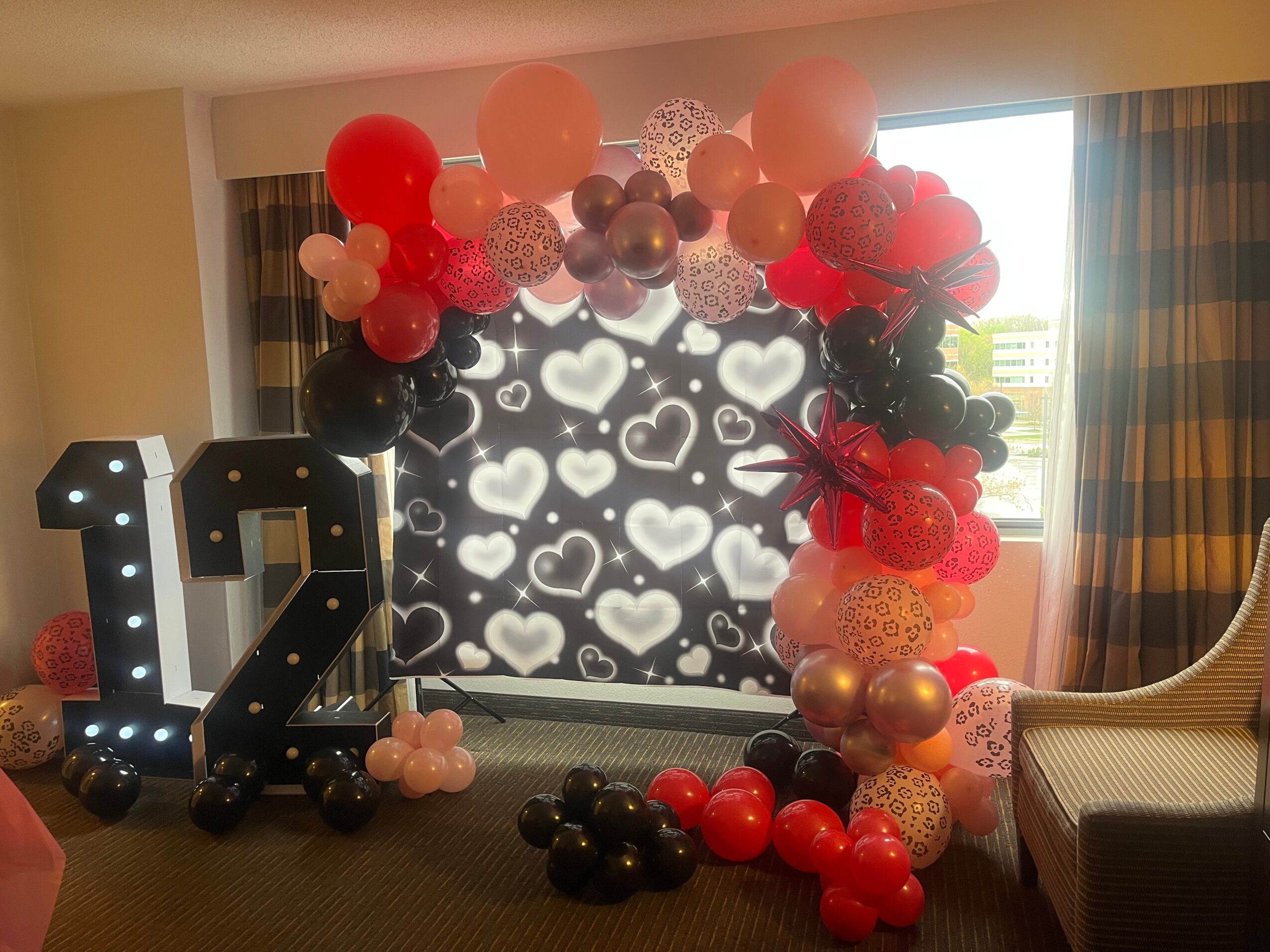 Decorative balloon arch with red, pink, black, and gold balloons surrounds a window with heart-shaped lights, and large illuminated number 12.