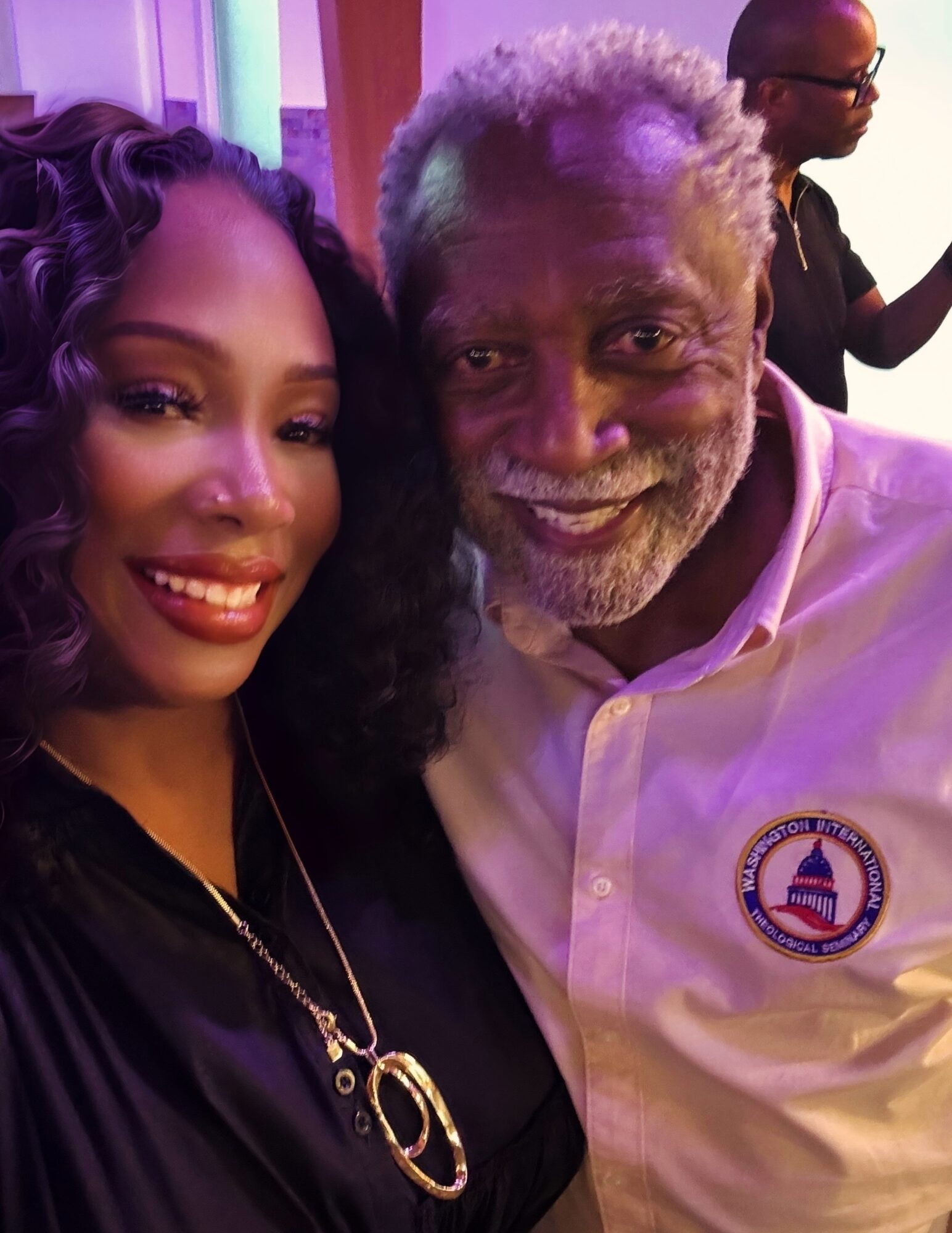 A woman and an older man smiling for a photo, woman with curly hair and dark clothing, man with gray hair and beard wearing a white shirt with a logo.