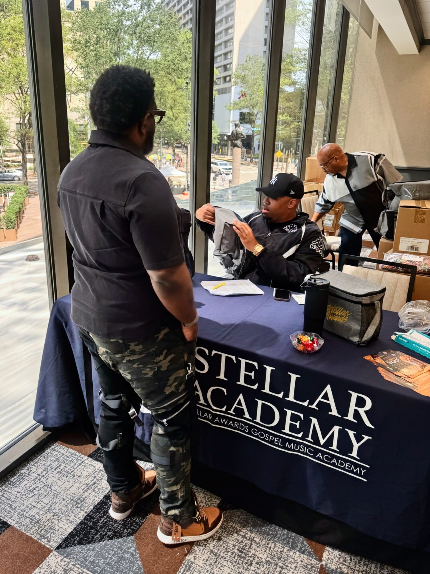 Person with glasses and curly hair standing at a table with a Stellar Academy banner, signing a book for a woman in black cap and jacket, in a bright indoor space.