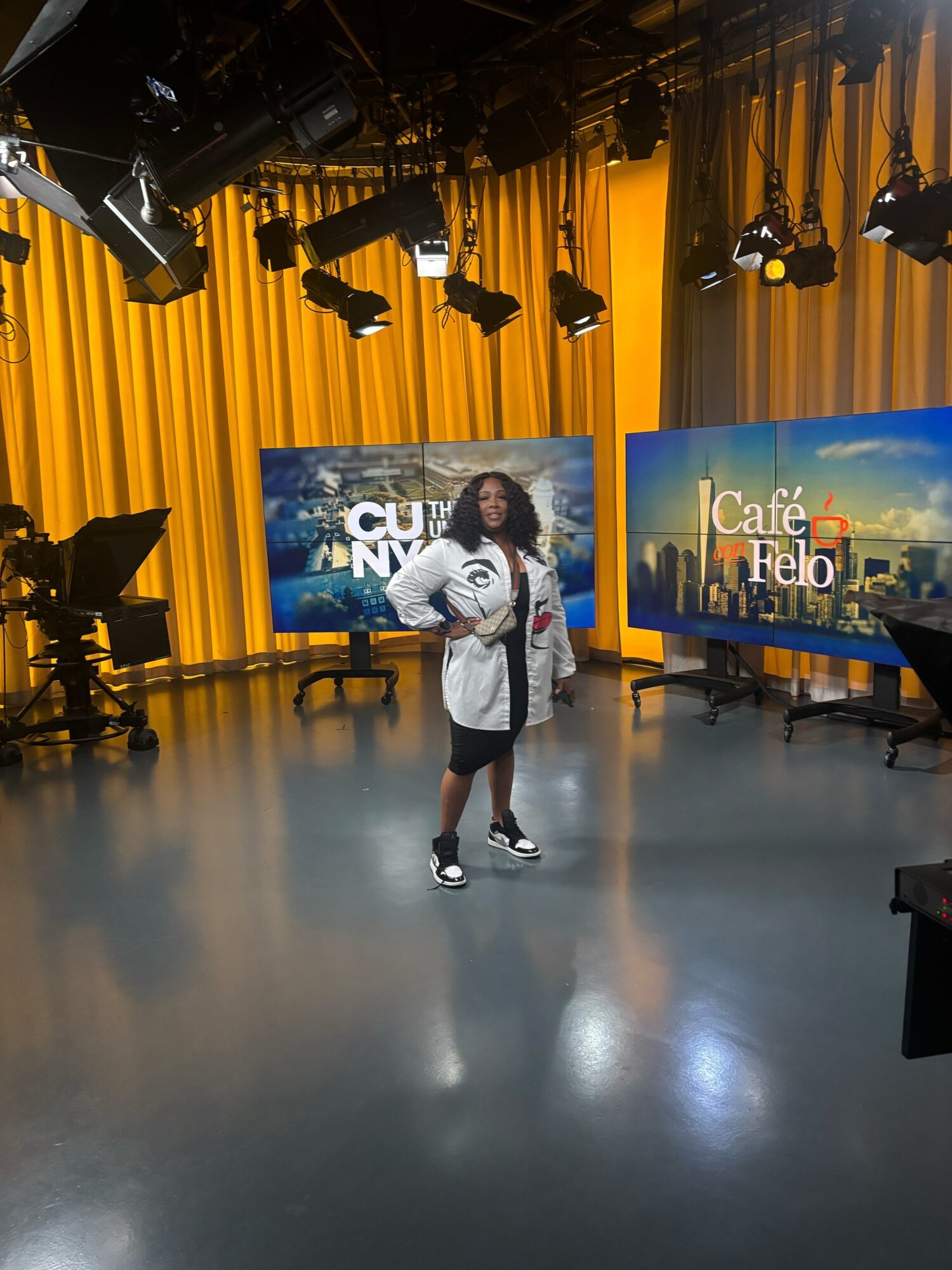 Person standing in a television studio with yellow curtains and large screens displaying cityscape images.