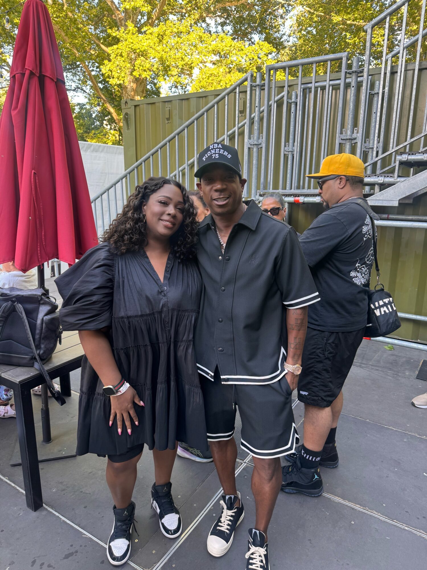 Two people standing outdoors, smiling, with a man wearing a black shirt and shorts, and a woman in a black dress, with a red umbrella and a metal fence in the background.