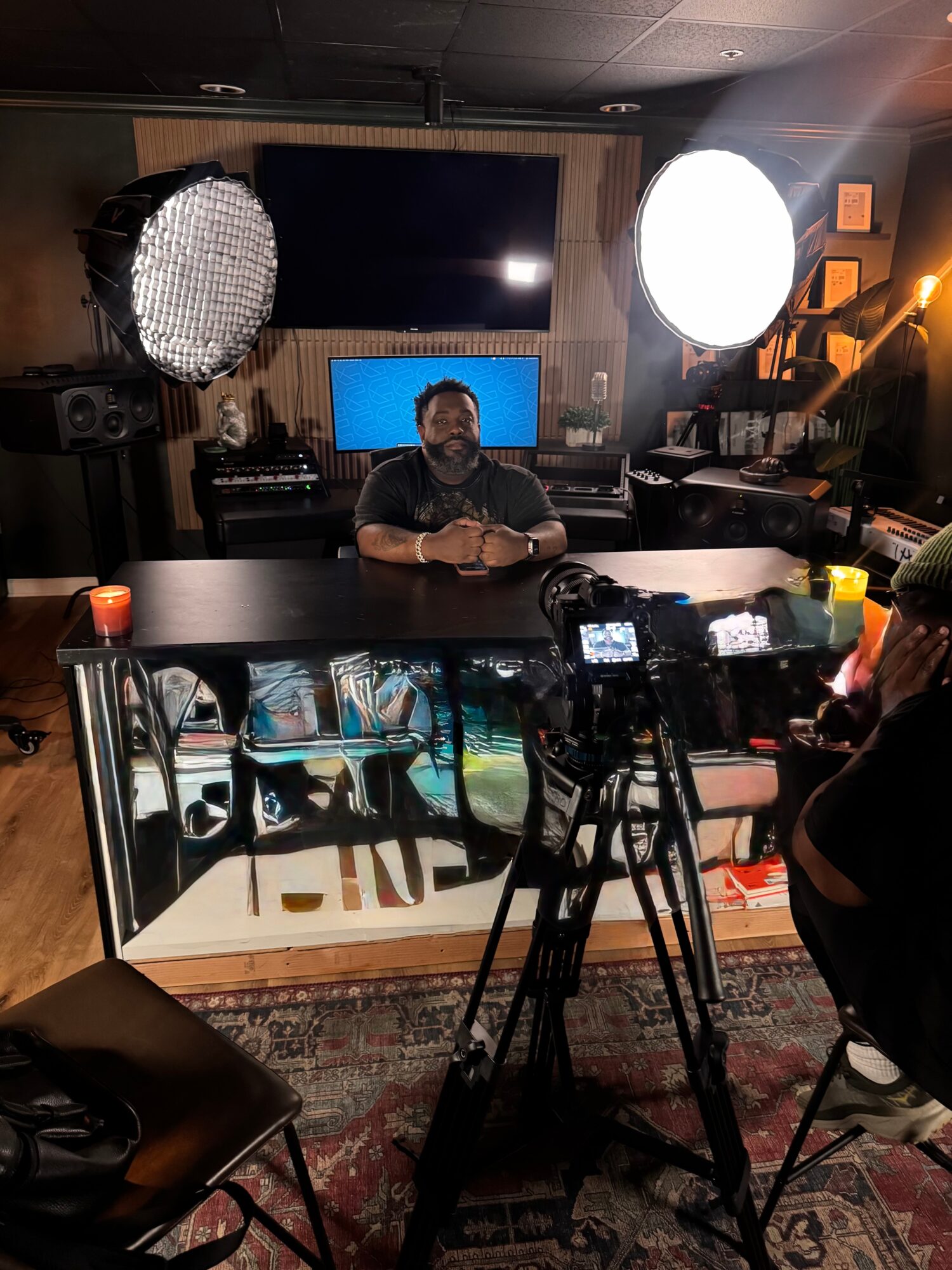 Person sitting at a desk in a recording studio with two large lights, a computer monitor, and audio equipment.