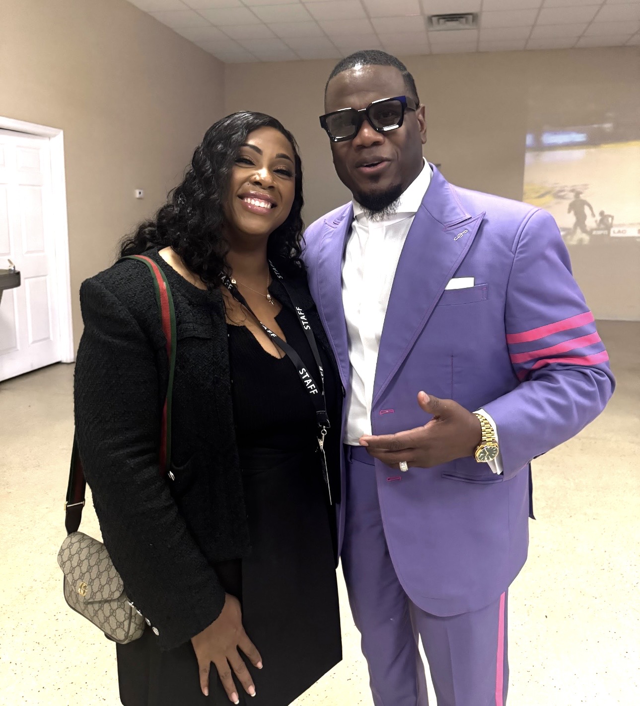 Two people standing indoors, smiling, with one wearing a black outfit and the other in a purple suit with sunglasses.