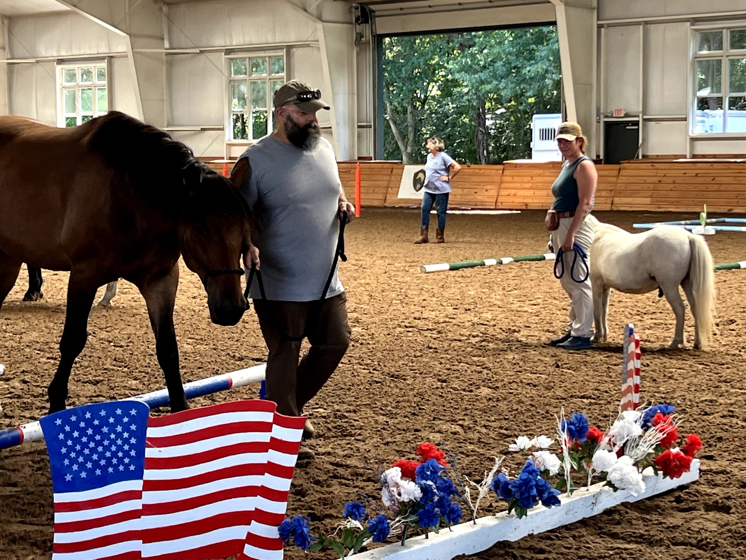People with horses inside an indoor riding arena, decorated with American flags and flowers, during a daytime event.