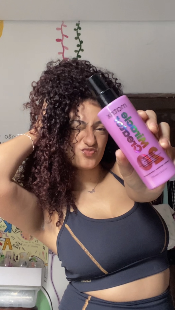 Woman with curly hair holding a pink bottle in front of her face, making a playful expression, in a room with colorful decorations
