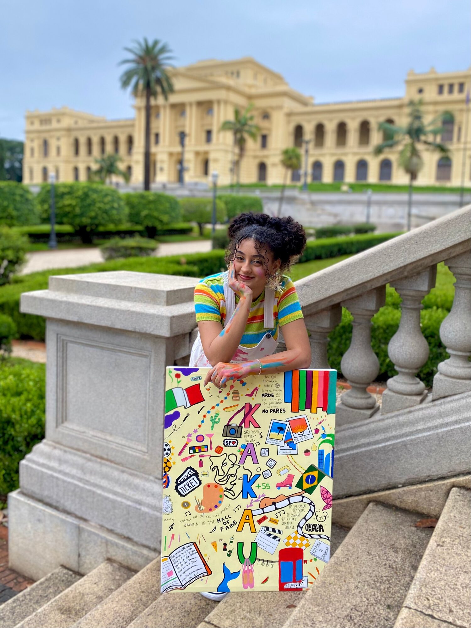 Child with curly hair in a colorful striped shirt leaning on a decorated board outdoors, with a historic building and palm trees in background.