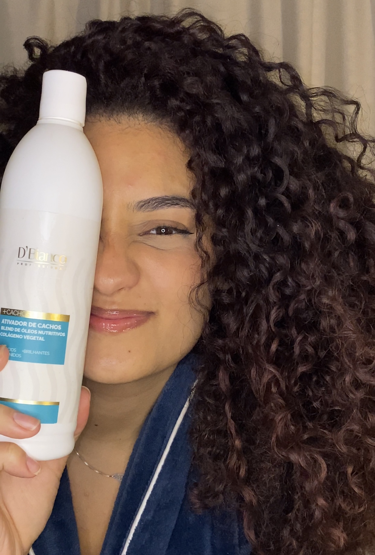 Person with curly hair holding a white bottle close to face, smiling with eyes partially closed.