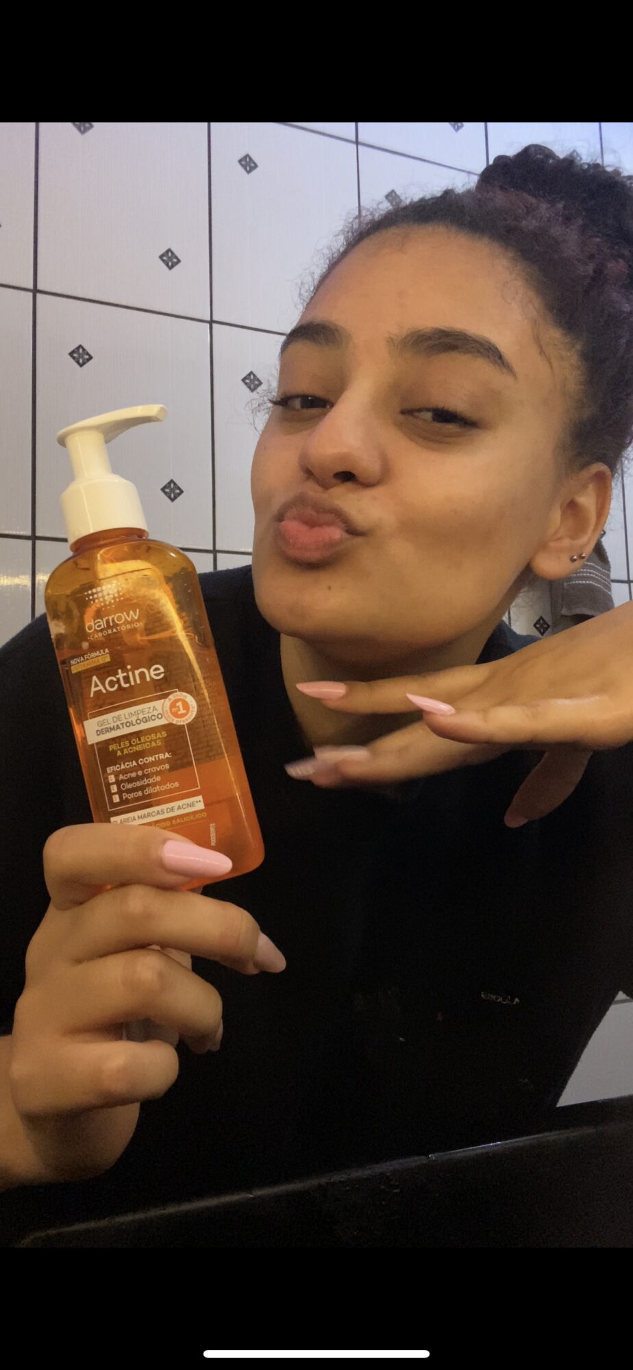 Young woman with dark hair in a bun holding a bottle of facial cleanser, making a duck face, in a tiled bathroom.