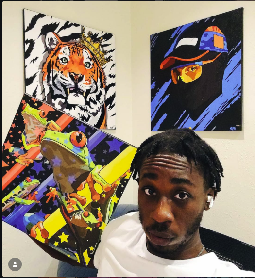Young man with dreadlocks in front of colorful animal and abstract paintings on a wall.