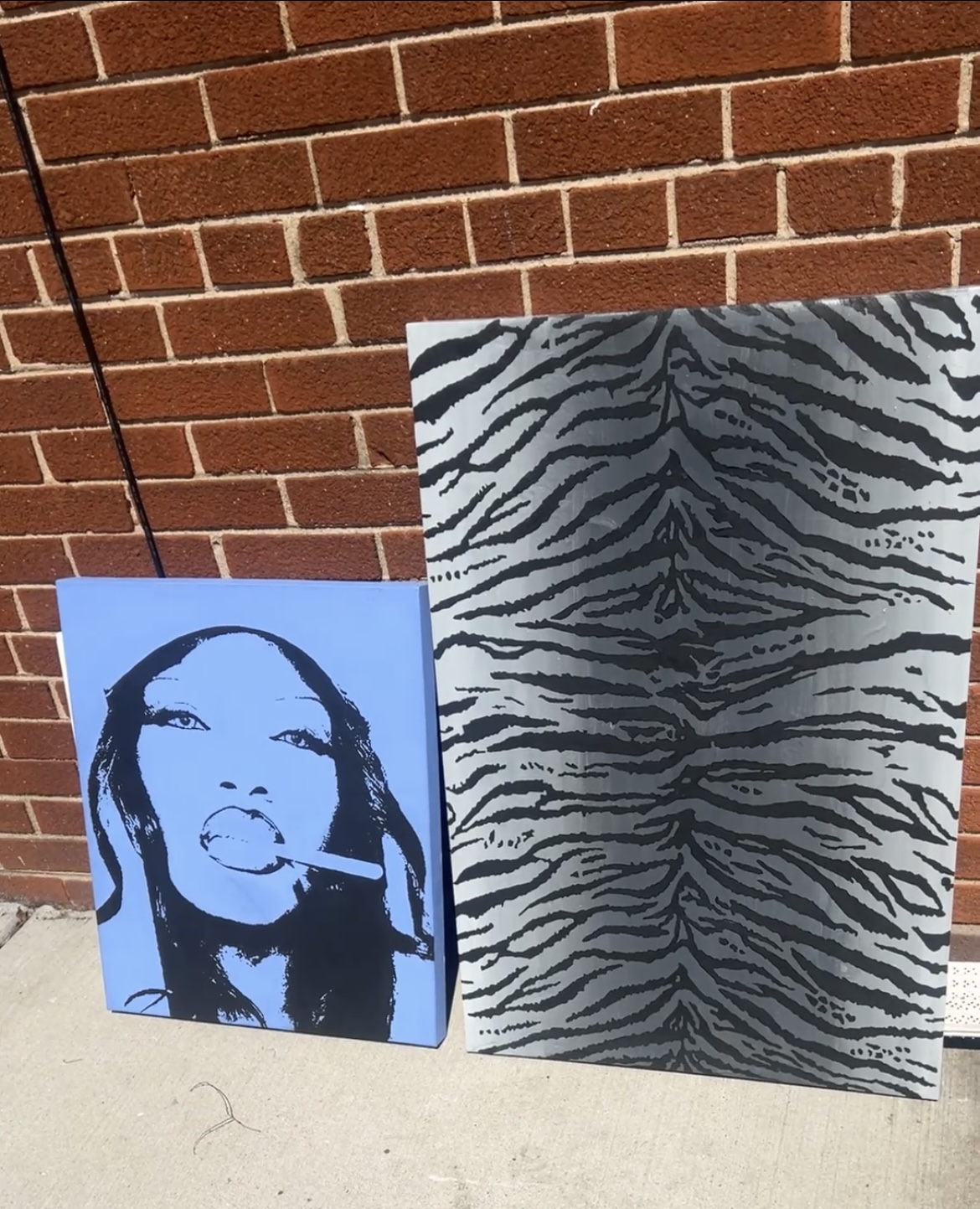 Two posters leaning against a brick wall, one with a stylized portrait of a woman and the other with a zebra pattern.