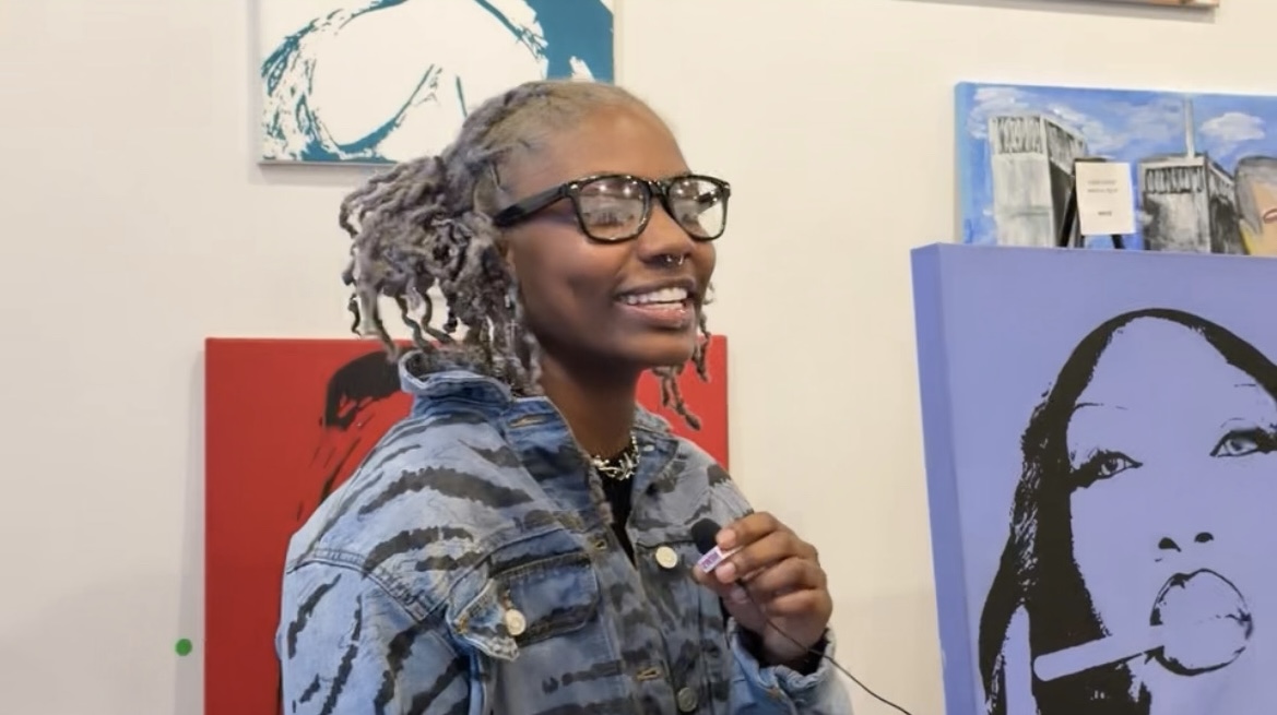 Smiling woman with glasses and gray hair styled in dreadlocks, wearing a denim jacket, standing in front of colorful artwork.