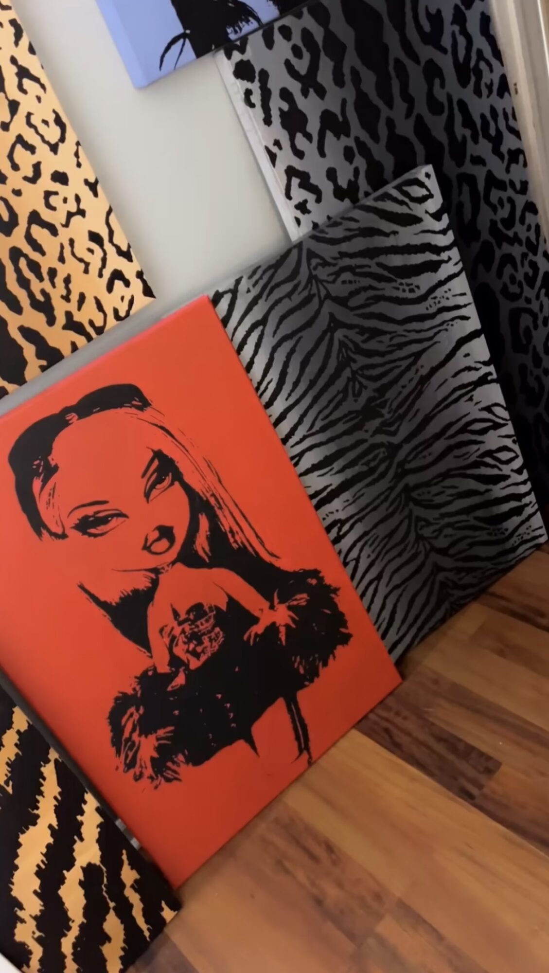 Three animal print patterned canvases leaning against a wall, with a portrait of a woman and dog in orange and black.