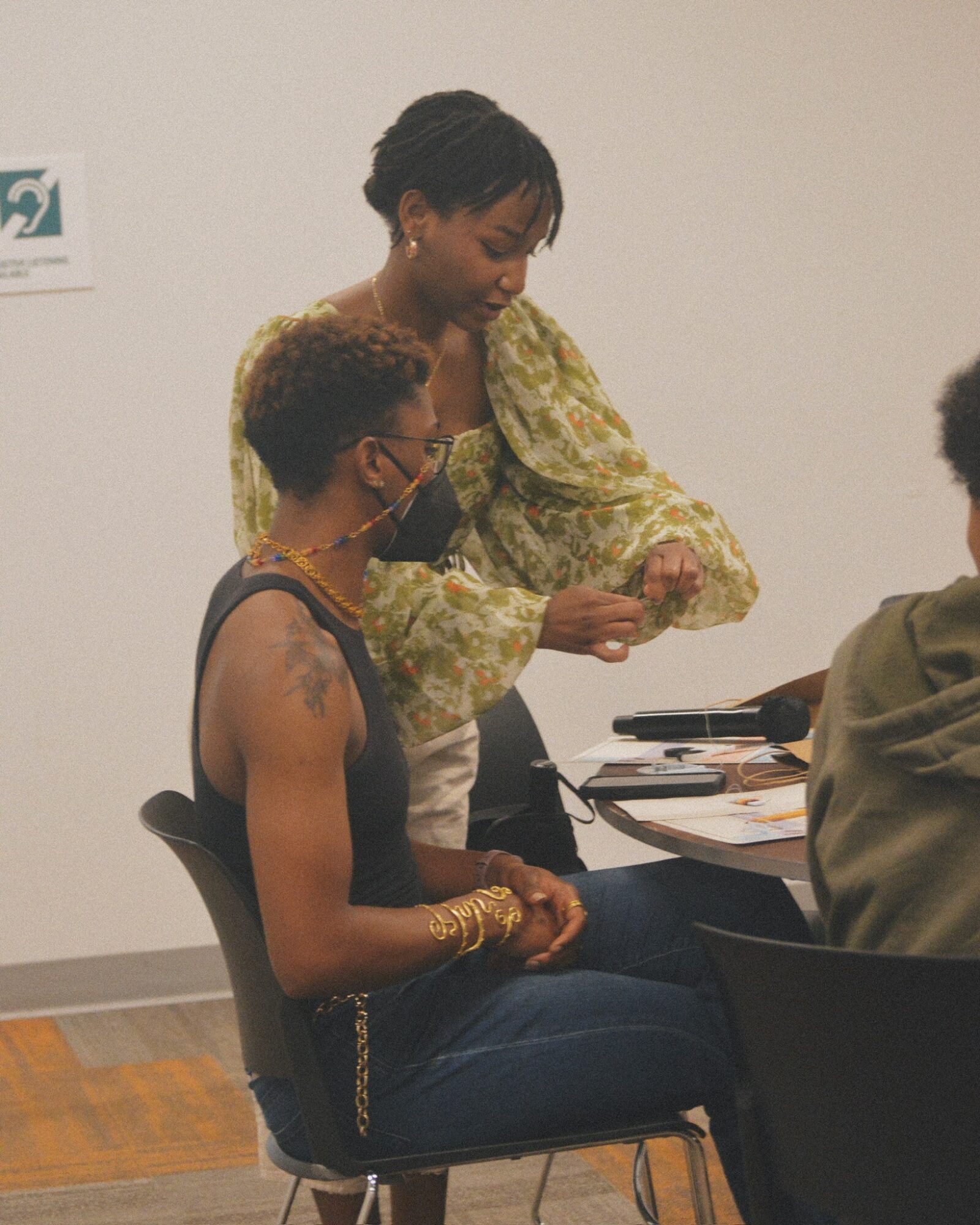 Two women sitting at a table, one standing and showing something to the other, in a room with a white wall.